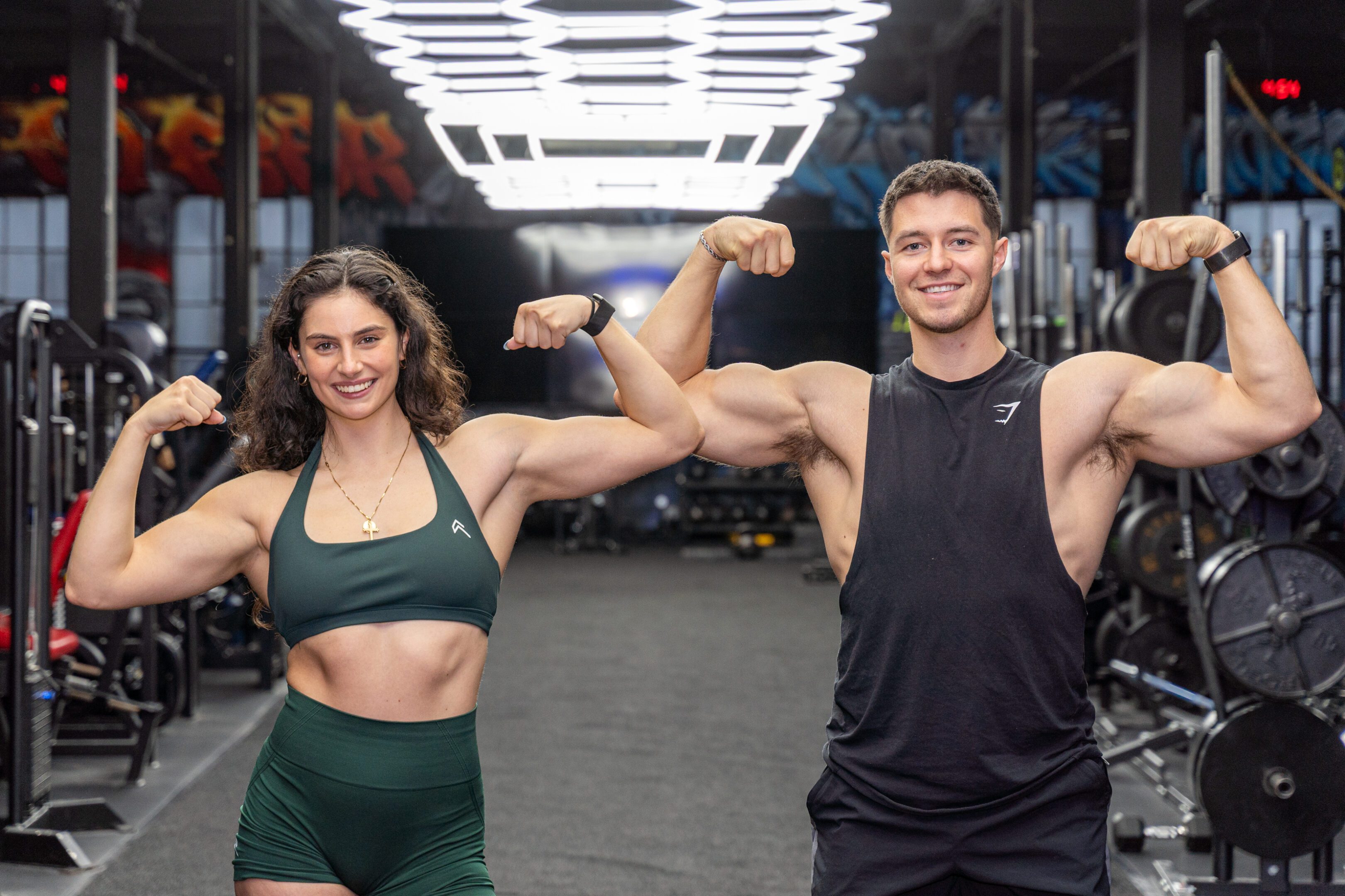 Two fit individuals flexing muscles and smiling in a gym.