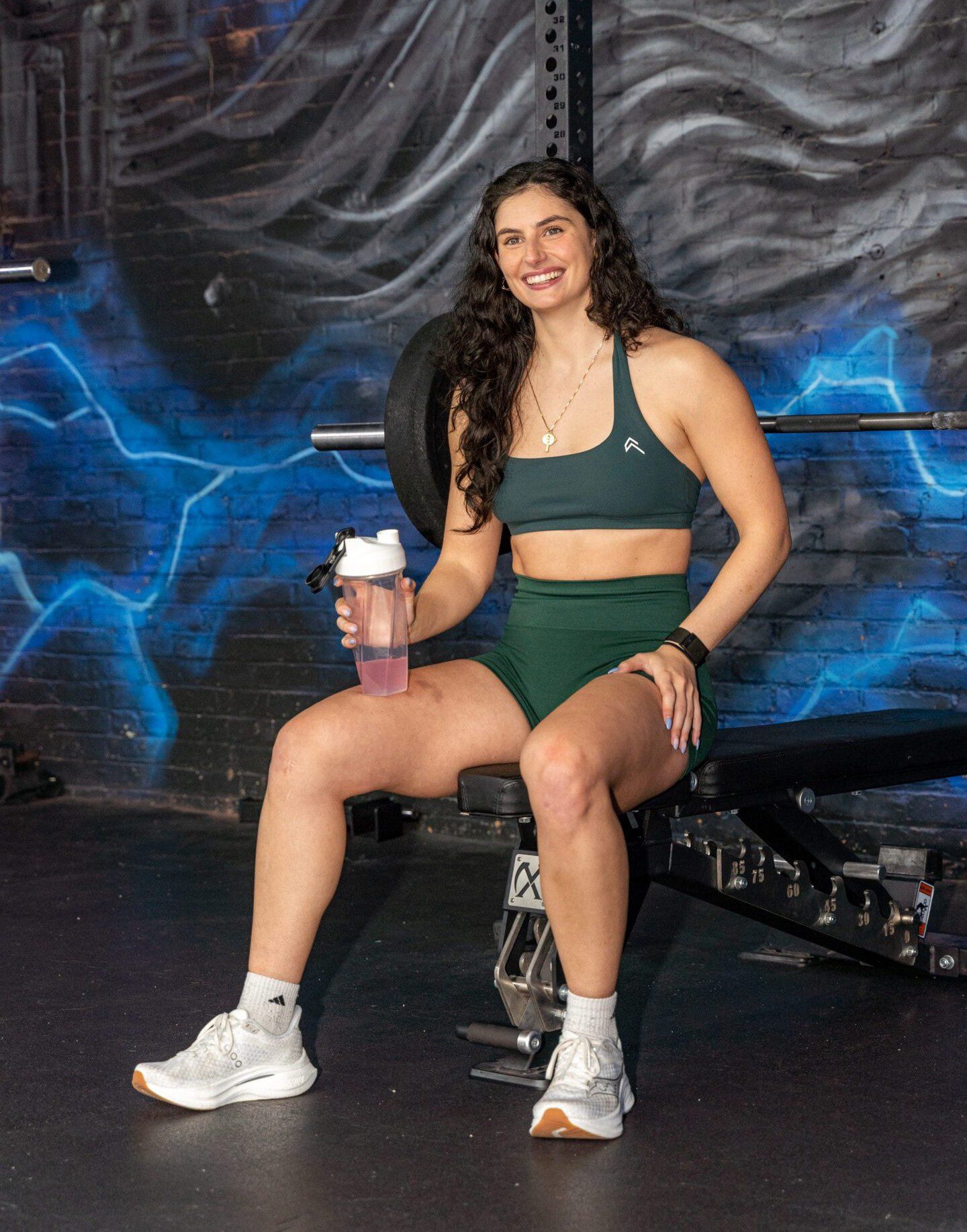 Woman in workout attire sitting on a bench holding a protein shake.