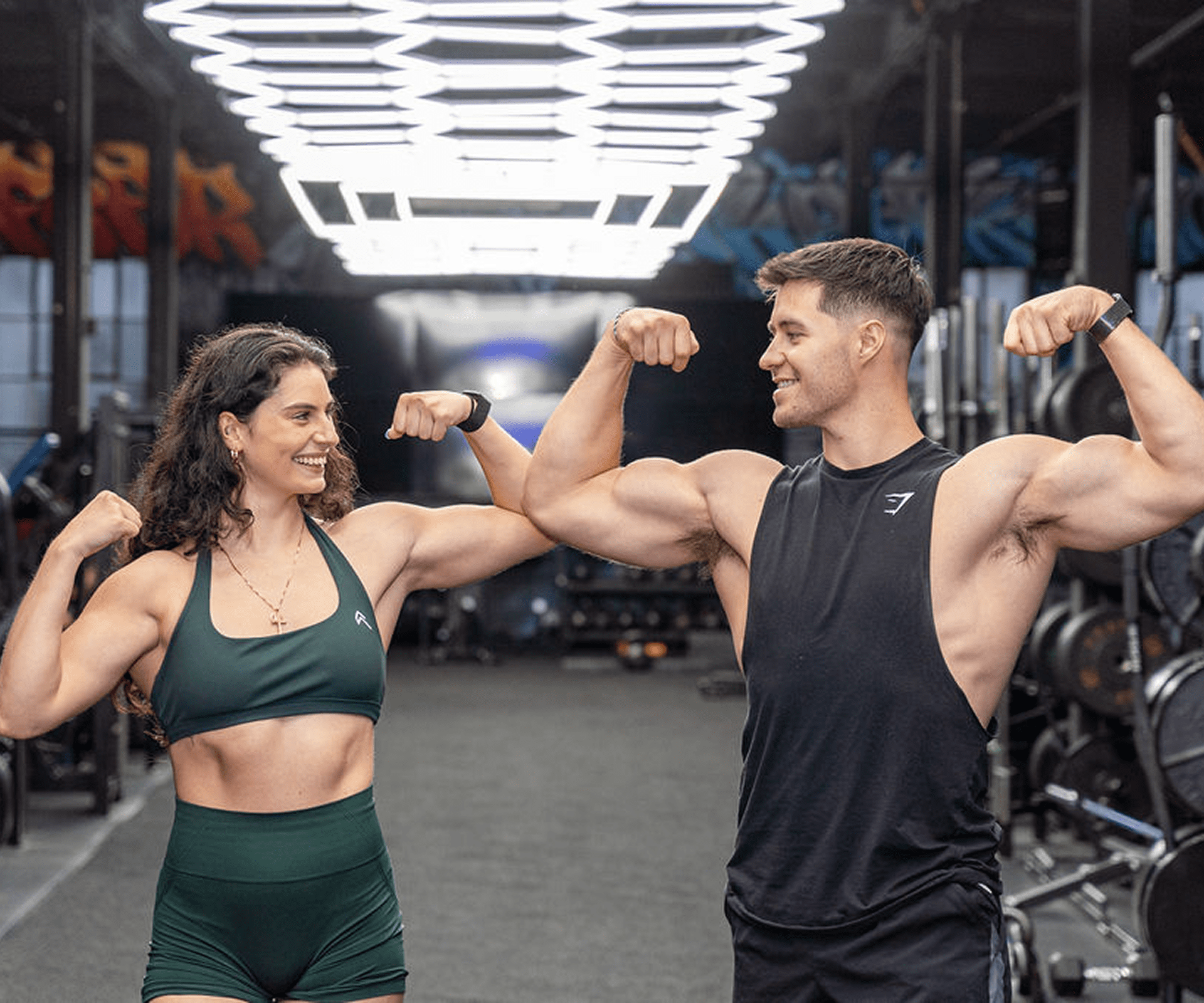 Two fit individuals flexing muscles in a gym, smiling at each other.